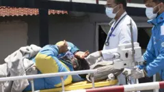 Dawa Sherpa levado em uma maca acompanhado por dois médicos. Ele veste uma jaqueta de montanhismo amarela e azul
