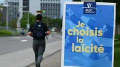 Homem caminha ao lado de um pôster de campanha política azul e amarelo, com os dizeres em francês 'Escolho o laicismo', em Longueuil, no Canadá