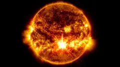 O Sol é visto como uma bola brilhante com redemoinhos claros e escuros; um grande clarão na superfície frontal representa uma poderosa erupção solar.