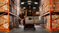 O fundador da empresa, Rubens Stuque, sentado em um sofá da marca erguido por uma pá carregadeira, em meio ao estoque da empresa