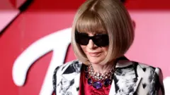 Anna Wintour, in her signature black sunglasses, poses on a red carpet in a red top and black-and-white patterned jacket