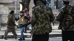 تصویر یک خبرنگار که به وسیله دو سرباز حکومت طالبان که یونیفرم نظامی بر تن و سلاح به دارند از کار بازداشته می شود.