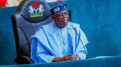 Aarẹ Bola Tinubu, o wọ agbada alawọ aro, o de fila alawọ aro, o lo igo si oju rẹ, kawọ le tabili, ẹrọ gbohungbohun wa niwaju rẹ, o n rẹrin-in musẹ