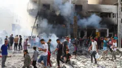 Crianças e adultos caminham sobre destroços em meio a fumaça