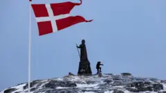 Umugabo aca iruhande y'ibendera rya Danemark hafi y'igishusho ca Hans Egede Statue i Nuuk