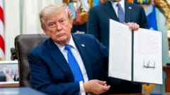 Trump, wearing a blue suit and sitting at desk in the Oval Office, holding up a signed executive order