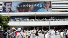 Fila de pessoas em frente a um estádio com um painel que tem a propagada do drama