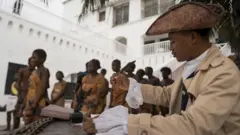 手前に18世紀ヨーロッパの服装を身にまとった人がいる。帽子をかぶり、机の上の文書に羽ペンで何か書こうとしている。奥では、薄い奴隷服を着た人々が鎖につながれて歩いている
