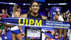 Farida Khalil, qui porte des lunettes transparentes et un gilet noir orné d'un dossard portant son nom de famille et le numéro un, tient une banderole sur la poitrine avec l'inscription Championnats du monde de pentathlon, Kaunas, Lituanie 2025. On peut voir plusieurs autres athlètes perplexe derrière elle dans un aréna intérieur, debout ou allongés sur le sol