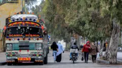 Mulheres caminham em rua movimentada de cidade síria, ao lado de ônibus decorado com lanternas e placas coloridas.