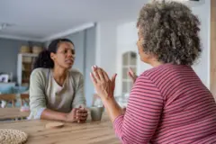 Dos mujeres sentadas a una mesa. Una mujer levanta sus manos para detener la conversación de la otra