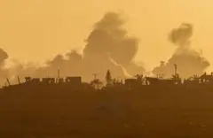 تصاعد الدخان من غزة بعد انفجار بالقرب من الحدود بين إسرائيل وغزة، كما شوهد من إسرائيل، الصورة تميل إلى اللون الأصفر. هناك القليل من الأشجار المتفرقة وبعض المباني صغيرة الحجم.
