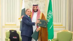 Suudi Arabistan Veliaht Prensi Muhammed bin Selman ve Pakistan Başbakanı Şehbaz Şerif ellerinde dosyalarla samimi bir şekilde gülümseyerek poz veriyor. Şerif'in bir eli bin Salman'ın koluna sarılmış, bin Salman'ın bir eli Şerif'in omuzunda. 