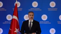 Hakan Fidan podyumda konuşuyor, arkasında Türk bayrağı ve mavi bir fon var 