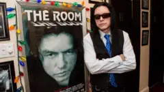 Tommy Wiseau com longos cabelos negros, óculos escuros e um colete preto encostado em um pôster de The Room, emoldurado por luzes coloridas, em uma exibição em Nova Orleans em 2014