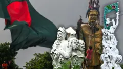 শেখ মুজিবুর রহমানের ভাস্কর্য ভাঙার দৃশ্য, ভেঙে ফেলা মুক্তিযুদ্ধের স্মরণে নির্মিত দুটি ভাস্কর্য এবং পেছনে বাংলাদেশের পতাকা 