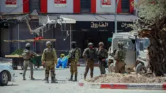 جنود إسرائيليون يقومون بدورية عند تقاطع طرق في مدينة جنين، الضفة الغربية، في 27 مايو/أيار 2025. اقتحمت قوات الاحتلال الإسرائيلي عدة مواقع، بما في ذلك محلات صرافة، وصادرت مبالغ مالية كبيرة، واعتقلت أفرادًا يُزعم ارتباطهم بفصائل المقاومة الفلسطينية. 