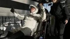 Butch erguendo os braços ao deixar cápsula, auxiliado por outras pessoas