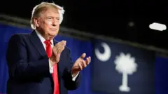 Perezida w'Amerika Donald Trump ahagaze ari ahavugirwa ijambo hegutse, yambaye ikositimu y'ubururu butsitse na karuvati itukura, arimo gukomera amashyi abantu batagaragara mu ifoto. 
