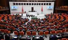 Turuncu renkteki Meclis sıraları yukarı bir açıdan görünüyor. Genel kurul salonunda kürsüde bir kadın var. Salonunu duvarlarındaki televizyonlarda konuşması yayınlanıyor.