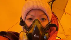Purnima está de pie dentro de una tienda de campaña de color amarillo brillante, vistiendo un traje de protección climática rojo para gran altitud y una máscara de oxígeno negra.