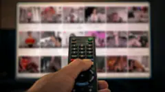 Uma mão mexendo em um controle de TV com uma TV ao fundo com imagens de pornografia