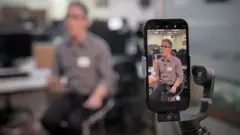 Romeu Zema, durante entrevista à BBC. O político aparece desfocado no segundo plano sentado em um banco alto, enquanto no primeiro plano vemos um celular filmando Zema, com a imagem dele em foco na tela