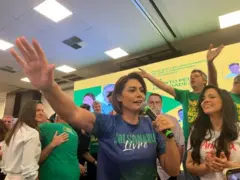Michelle Bolsonaro falando em um microfone com uma das mãos erguidas. Ela usa uma blusa que se lê: Bolsonaro livre