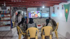 Cinco soldadas sentam-se em cadeiras de plástico e assistem à televisão em uma sala improvisada em uma caverna. Elas vestem uniformes cáqui e estão de costas para a câmera.