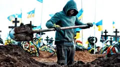 Muškarac kopa grob na groblju u Odesi 16. marta 2024. Oko deset ukrajinskih zastava vijori se na belim jarbolima iza njih. Kopač groba u nosi tirkiznu duksericu sa kapuljačom, u rukama mu je lopata