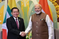 ဗိုလ်ချုပ်မှူးကြီး မင်းအောင်လှိုင်နဲ့ အိန္ဒိယ ဝန်ကြီးချုပ် မိုဒီတို့ လက်ဆွဲနှုတ်ဆက်နေကြ 
နှစ်ယောက်စလုံးက ပြုံးပြီး ကင်မရာကိုကြည့်နေ။ မြန်မာစစ်ခေါင်းဆောင်ဟာ အနောက်တိုင်း ဝတ်စုံကို ဝတ်ထားပြီး အိန္ဒိယဝန်ကြီးချုပ်ကတော့ အညိုရောင်အပေါ်ထပ်  အိန္ဒိယ ရိုးရာထည်ကို အဖြူရောင် ရှပ်အင်္ကျီ လက်ရှည်နဲ့အတူ ဝတ်ထားတာတွေ့ရပါတယ်။ သူတို့နှစ်ဦးအနောက်မှာတော့ အိန္ဒိယနဲ့ မြန်မာ အလံတွေ ရှိနေ 