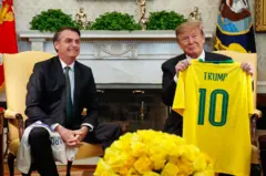 Foto de Bolsonaro e Trump em sala da Casa Branca. Os dois estão sentados em e Trump exibe uma camiseta do Brasil com o nome Trump nas costas