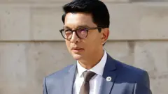 Andry Rajoelina porte un costume bleu, une chemise blanche, une cravate sombre et des lunettes.