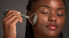 Mulher negra utiliza rolo de jade para massagem facial em estúdio, sobre um fundo cinza.