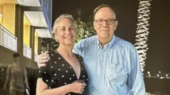 Gina Montaner y su padre, Carlos Alberto Montaner, en Miami con un apartamento al fondo y el tronco de una palmera iluminado. El padre rodea con el brazo a su hija, que sonríe.