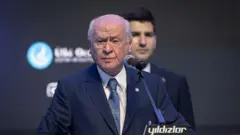 Bahçeli kürsüde lacıvert takım elbisesiyle ayakta konuşma yapıyor. Beyaz seyrek saçları ile sert bir duruşu var. Arkada bir erkek daha duruyor.