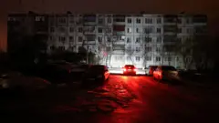 На заднем плане - обесточенный панельный пятиэтаный многоквартиный дом. На переднем плане дорога, освещенная красными фарами автомобиля перед домом.