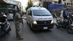 Uma ambulância passa por uma rua movimentada, passando por um homem em uniforme militar com um rifle de assalto