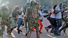 Abigaragambya ku nteko ishingamategeko bariruka banyura ku bapolisi bazamuye inkoni, mu myigaragambyo y'i Nairobi, muri Kenya, ku itariki ya 25 Kamena (6) mu 2024
