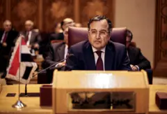  فهمي يرتدي بدلة رسمية تبدو كحلية اللون وقميصاً أبيض وربطة عنق حمراء ونظارات طبية، بشرته تميل إلى السمرة. يبدو كبيراً في السن لكن شعره لونه أسود. يجلس على طاولة اجتماعات وكتب أمامه (جمهورية مصر العربية) وخلفه عدد من الأشخاص.