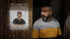 Jads Vater steht neben einem Plakat zum Gedenken an seinen Sohn, das an der geschnitzten Holzeingangstür der Familie im Flüchtlingslager al-Far'a im besetzten Westjordanland angebracht ist.
