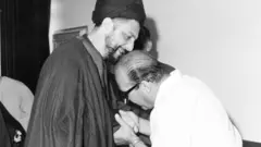 Sadr, sonriente, se inclina hacia un hombre con anteojos y vestido de blanco que sostiene las manos de Sadr para besarlas