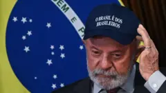 O presidente Lula com um boné escrito "o Brasil é dos brasileiros" e a bandeira do Brasil no fundo.