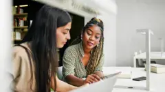 Jovem explica algo a outra, com as duas em frente a um computador