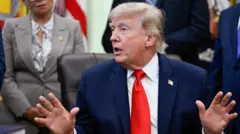 Perezida w'Amerika Donald Trump yicaye mu ntebe y'imisego arimo kuvugira mu biro bye bya White House i Washington DC, muri Amerika. Akoze ikimenyetso n'ibiganza, inyuma ye hahagaze umugore umwe n'abagabo babiri bambaye amakositimu, bataboneka mu maso. Ifoto yo ku itariki ya 11 Kamena (6) mu mwaka wa 2026.