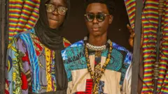 Deux hommes vêtus de vêtements patchwork colorés à motifs multiples regardent la caméra encadrée par une porte, à Diourbel, au Sénégal, en avril 2024. 