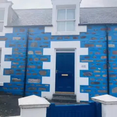 Uma casa de tijolos pintada de azul e branco. A porta é azul e ao lado está o número 115