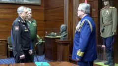 Начальник ГРУ адмирал Игорь Костюков в черной форме, еще один офицер в российской зеленой форме, офицер в иностранной синей форме стоят напротив друг друга среди стеллажей с макетом танка, манекеном в солдатской форме и бюстом