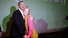 Кандидатка от Партии зеленых Ханна Спенсер и лидер партии Зак Полански стоят, обнявшись, на фоне логотипа своей партии и радостно улыбаются