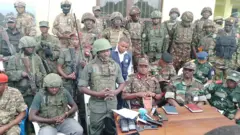 Col Imani Nzenze wa M23 (uhagaze imbere) mu muhango wo gushyikiriza Kibumba, agace kari kuri 25km uvuye i Goma, ingabo z'akarere zizaharinda 